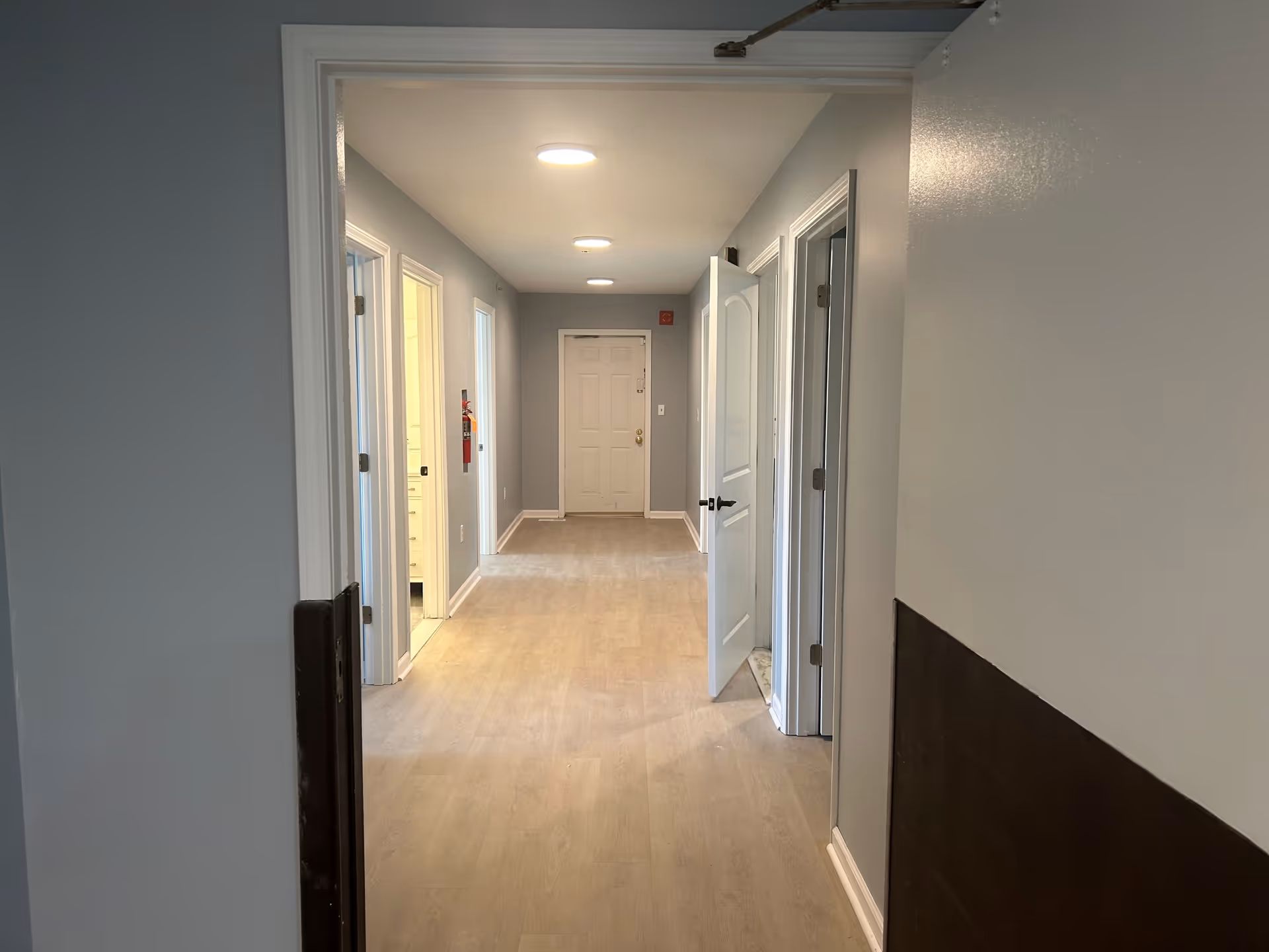 A clean interior hallway with light wood flooring, gray walls, recessed ceiling lights, and several open doors leading to rooms.