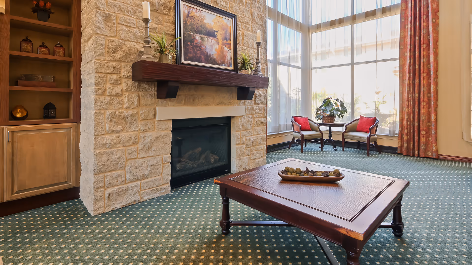 A cozy living room area with a stone fireplace, a wooden mantel decorated with candles and plants, a framed painting above the mantel, a wooden coffee table with a decorative bowl, two armchairs with red cushions, a small round table with a potted plant, large windows with sheer curtains, and patterned green carpet.