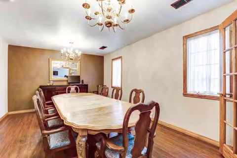 A dining room with a long wooden table surrounded by eight wooden chairs with cushioned seats. The room has hardwood flooring, two windows with wooden frames, a chandelier hanging from the ceiling, and a sideboard with a large mirror above it against the far wall.