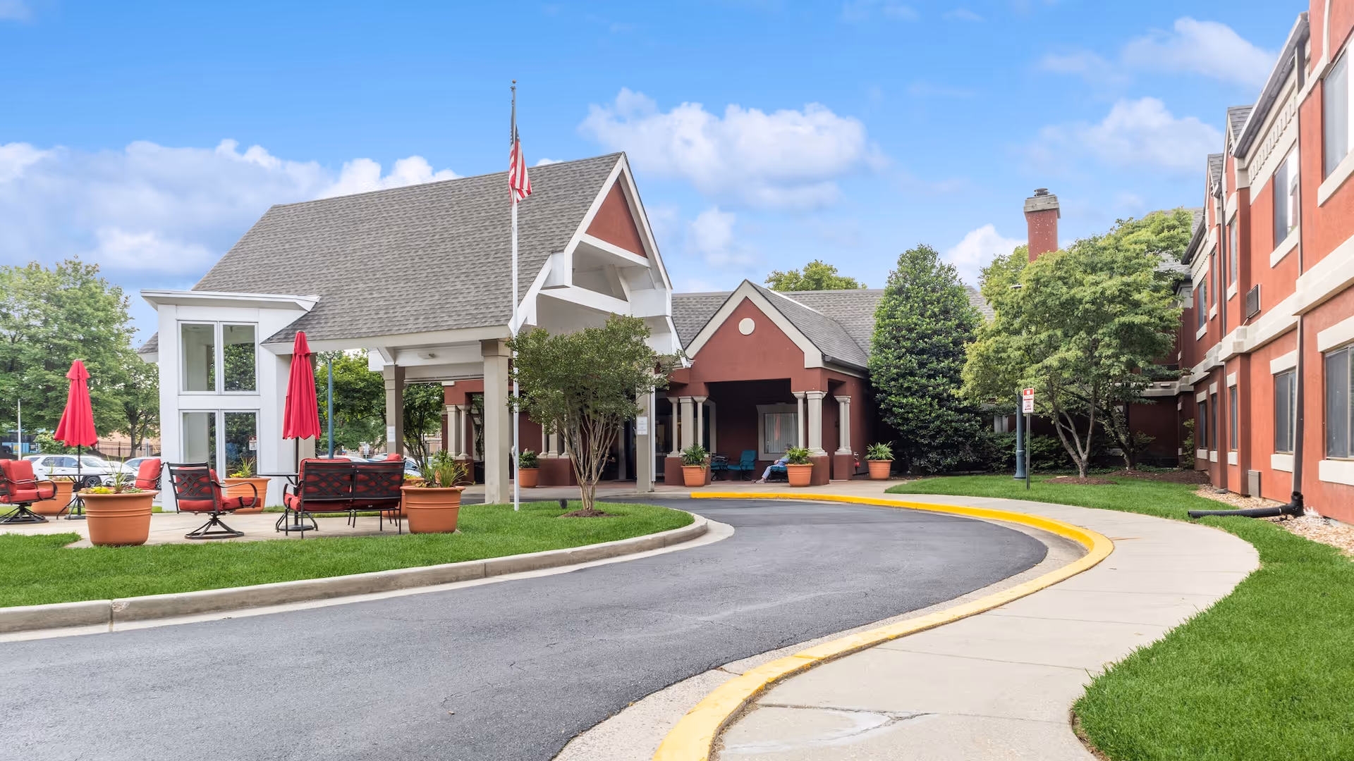 Front exterior view of Aspenwood Senior Living Community featuring a covered entrance with a flagpole, outdoor seating with red umbrellas, and a curved driveway surrounded by green grass and trees under a partly cloudy sky.