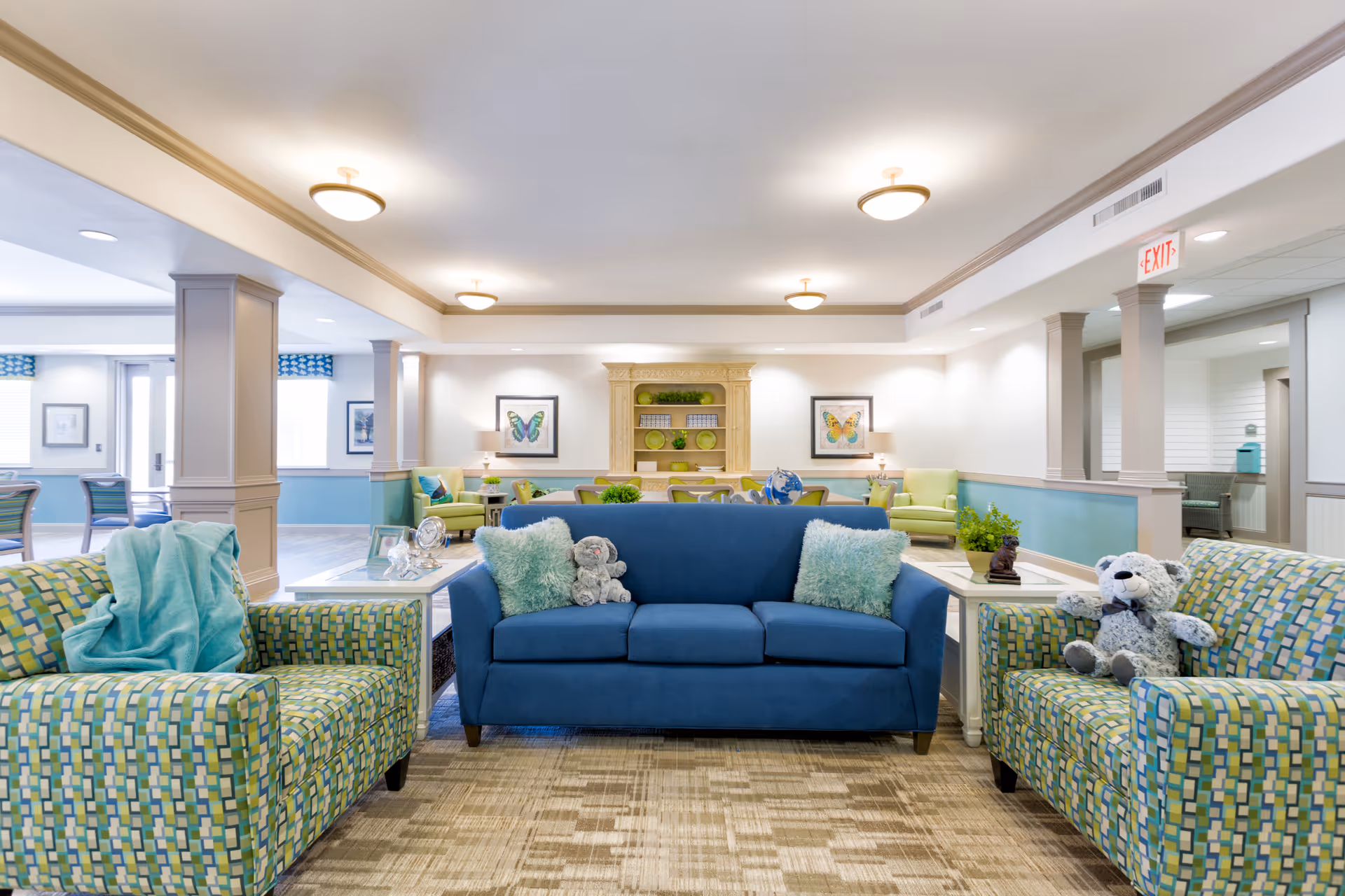 A bright and spacious senior living common area with a blue sofa and two patterned armchairs, each adorned with plush pillows and stuffed animals. The room features soft lighting, framed butterfly artwork on the walls, and a decorative wooden shelving unit with green accents. The space has a welcoming and cozy atmosphere with light-colored walls and carpeted flooring.
