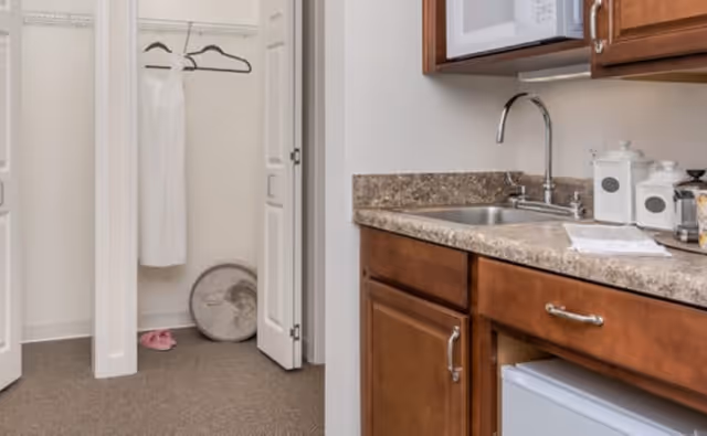 Interior view of a small kitchen area with a sink, countertop, wooden cabinets, and a microwave above. To the left, there is an open closet with a white dress hanging on a hanger and a pair of pink slippers on the floor.