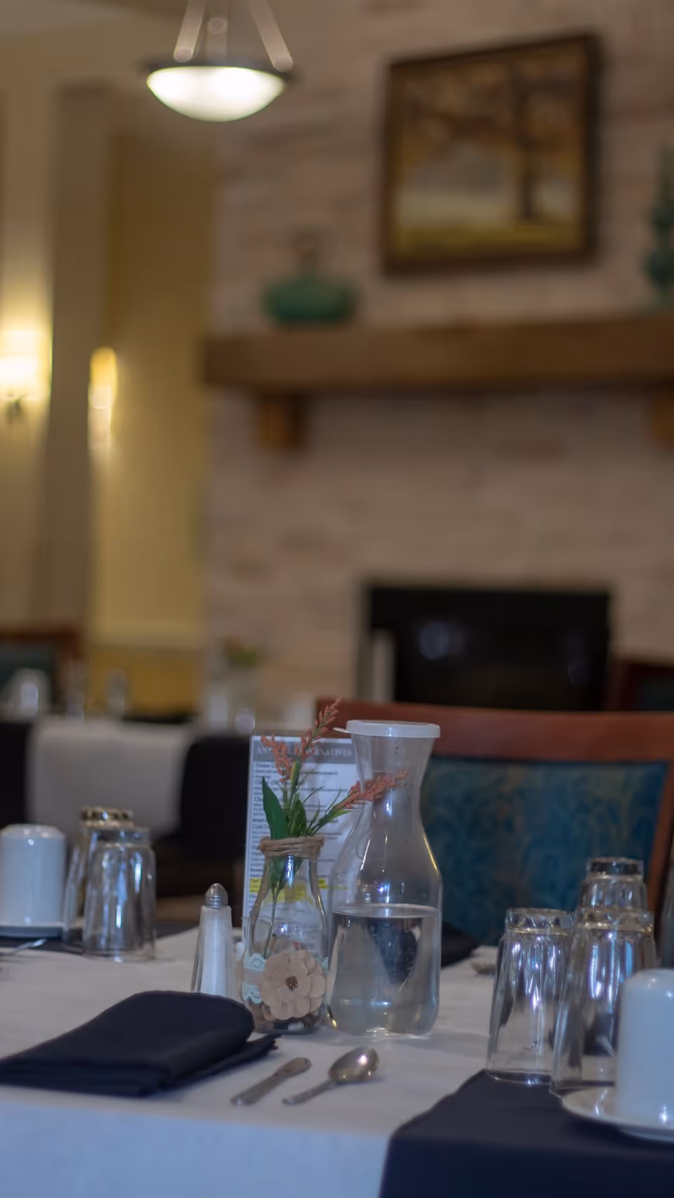 A dining table set with a water carafe, a small decorative vase with flowers, salt shaker, utensils, and upside-down drinking glasses in a cozy dining room with a fireplace and framed artwork in the background.