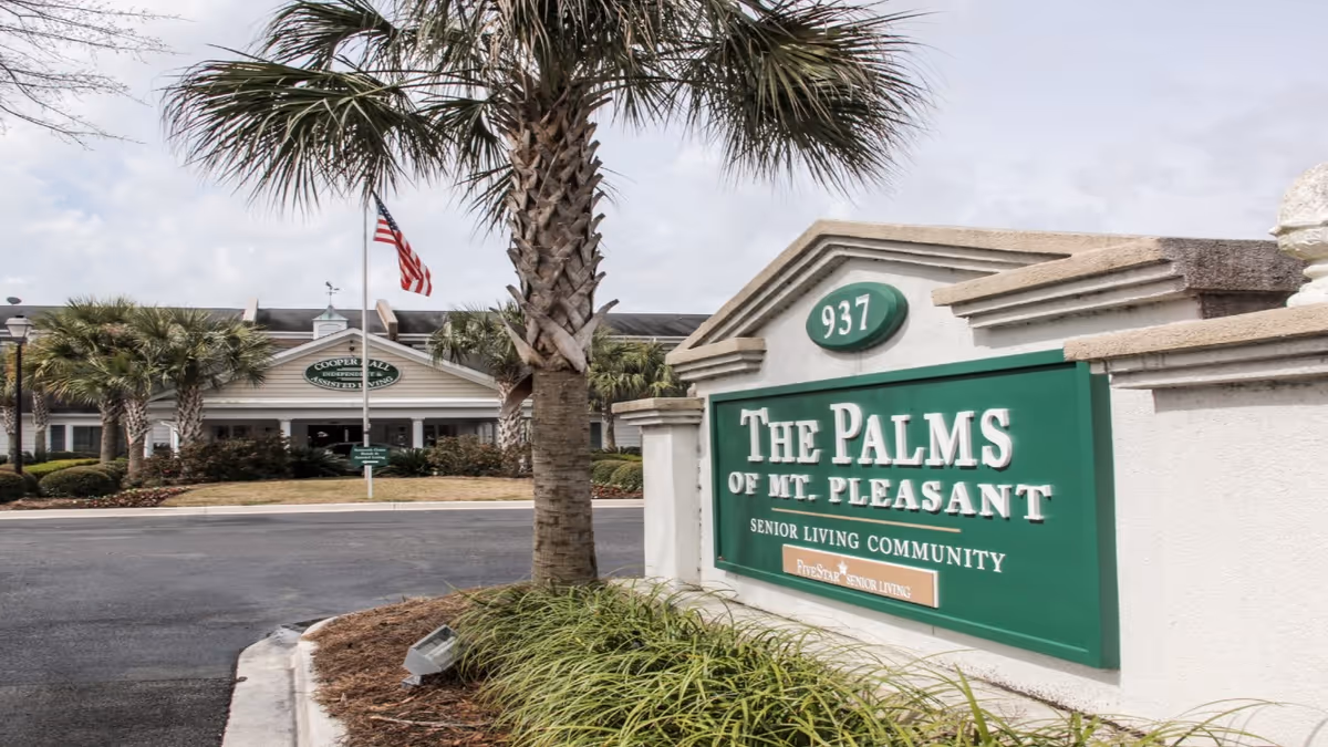 Entrance sign for The Palms of Mt. Pleasant senior living community with palm trees and the community building in the background under a cloudy sky.