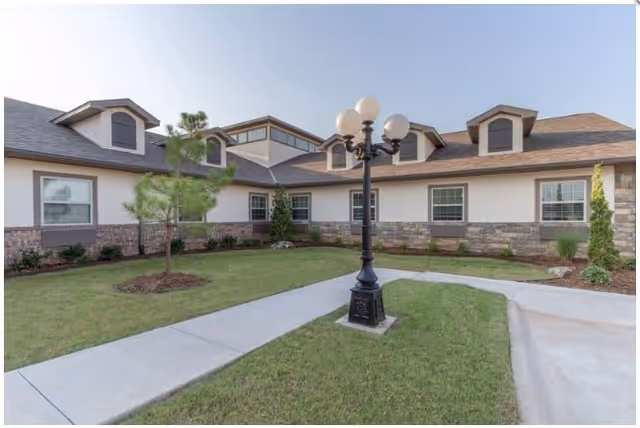 Exterior view of a single-story building with beige walls and stone accents, surrounded by a well-maintained lawn and small trees. A black lamp post with multiple white globes stands on a concrete walkway leading to the building entrance under a clear blue sky.