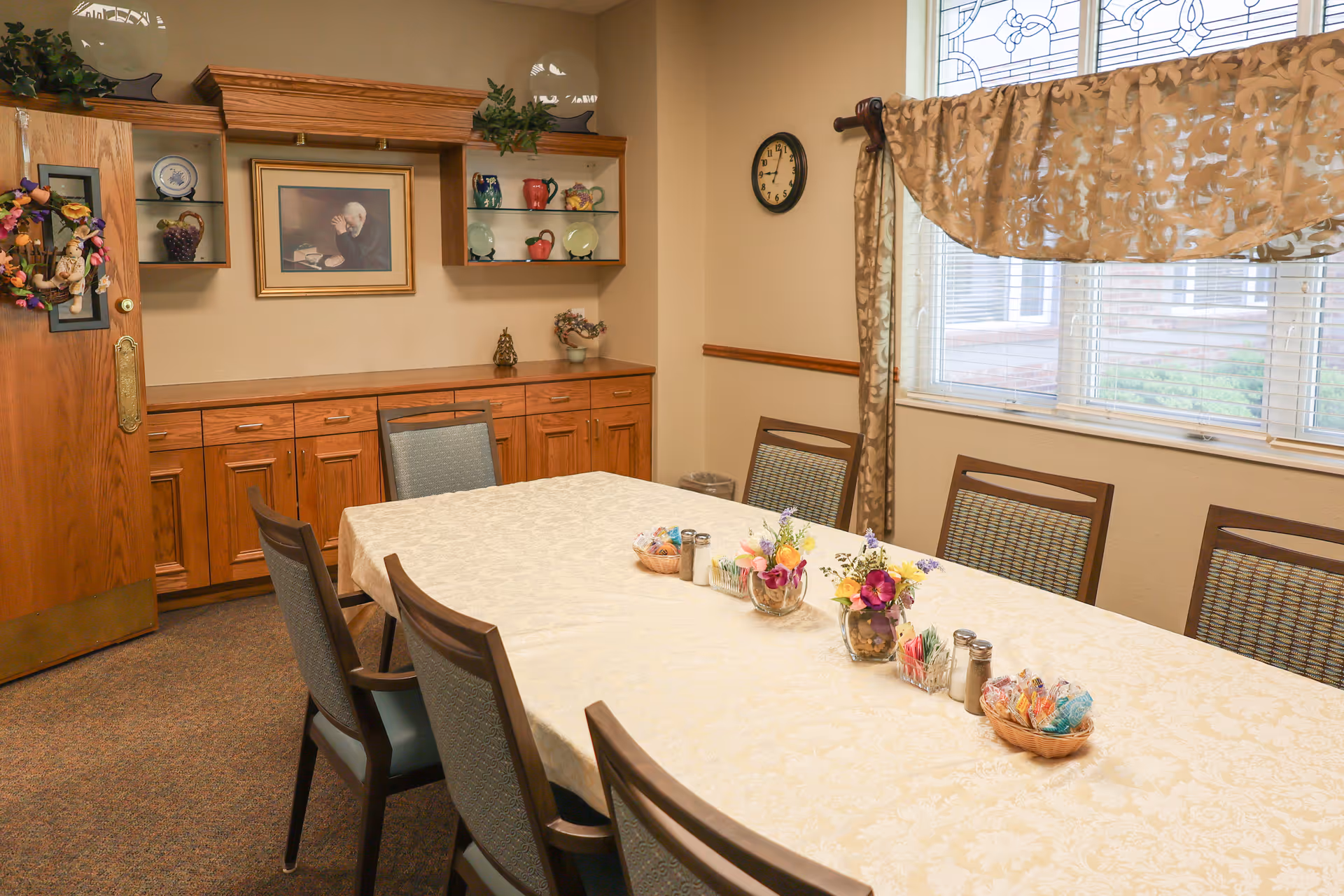 A dining room with a long table covered with a cream-colored tablecloth, surrounded by eight chairs. The table is decorated with small flower arrangements, salt and pepper shakers, and a basket of wrapped candies. There is a wooden cabinet with glass shelves displaying decorative plates and pitchers, a framed picture on the wall, a clock, and a window with patterned curtains letting in natural light.