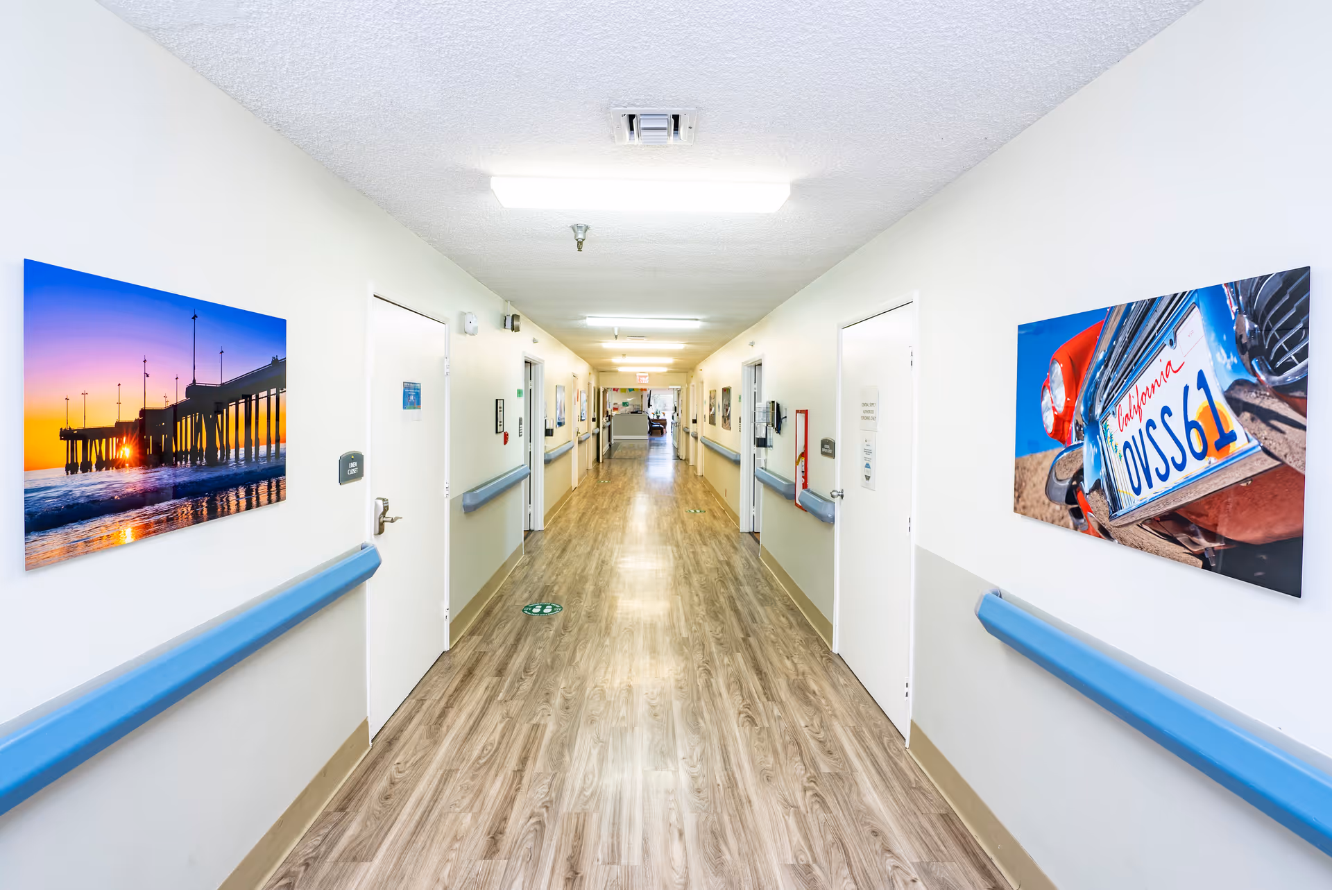 A long, clean hallway in a care facility with light-colored walls and wood-patterned flooring. Blue handrails run along both sides of the hallway. Several closed white doors line the corridor, and colorful framed photographs hang on the walls. The ceiling has fluorescent lighting fixtures.