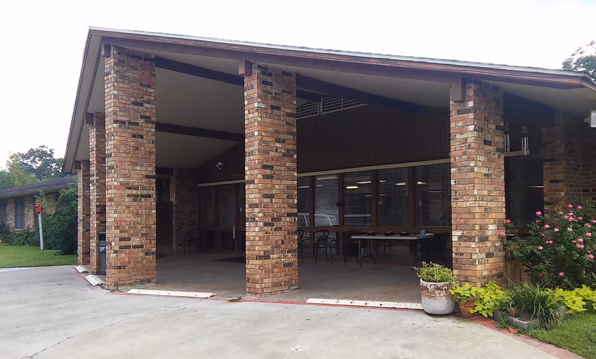 Exterior view of a brick building with a covered patio area supported by brick columns. There are tables and chairs under the patio, potted plants near the columns, and greenery surrounding the building.