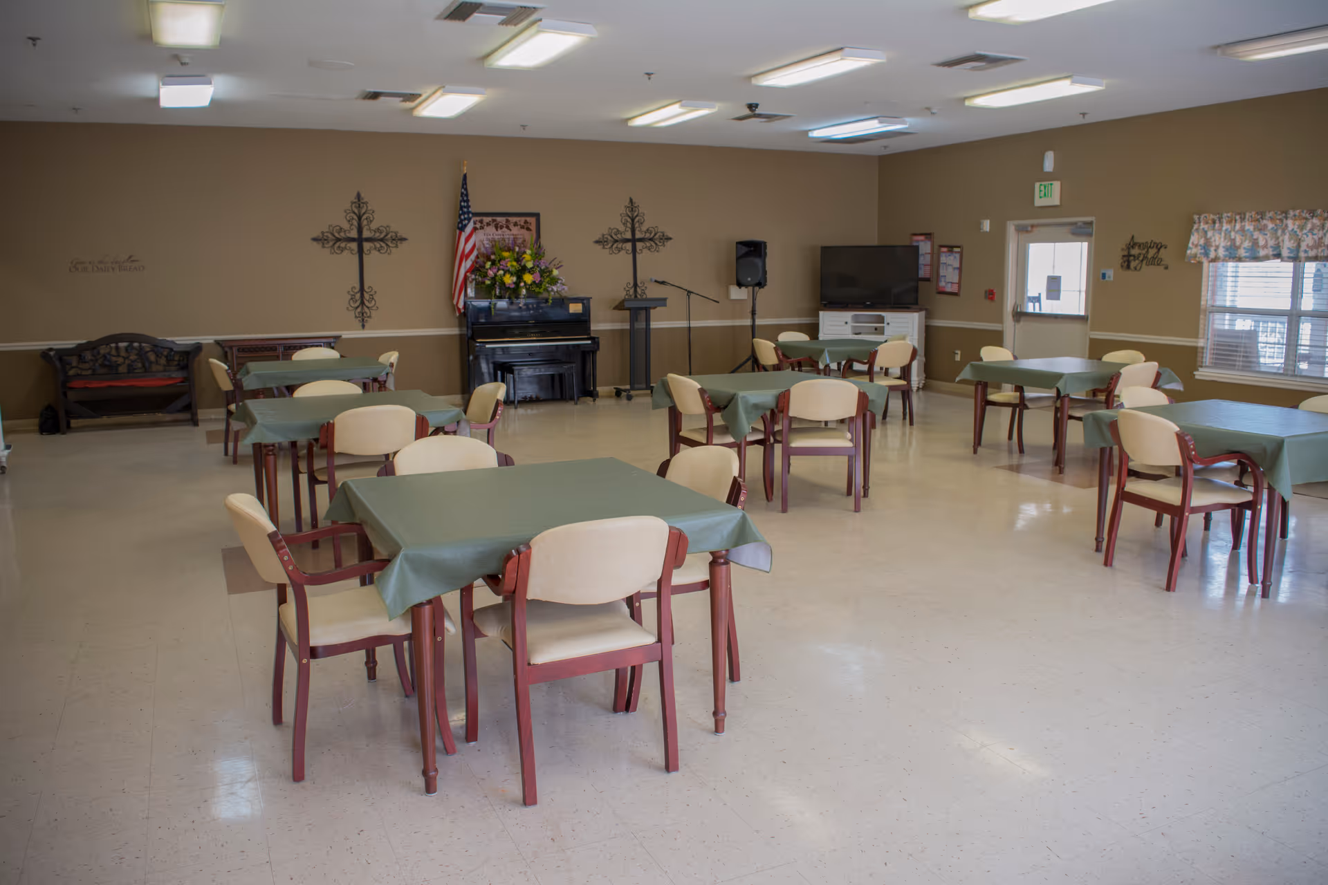 A spacious dining room with several tables covered with green tablecloths and surrounded by beige chairs with wooden frames. The room has beige walls decorated with two large crosses, an American flag, a piano with a flower arrangement on top, a podium, a speaker, and a television on a white stand. There is a window with floral curtains and a door with an exit sign above it.