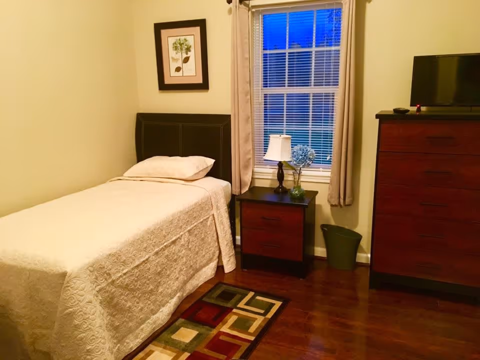A small bedroom with a single bed covered in a cream-colored quilt. Next to the bed is a wooden nightstand with two drawers, a table lamp, and a vase with blue flowers. A window with beige curtains is behind the nightstand. On the right side, there is a tall wooden dresser with a flat-screen TV on top. A colorful geometric-patterned rug is partially visible on the wooden floor.