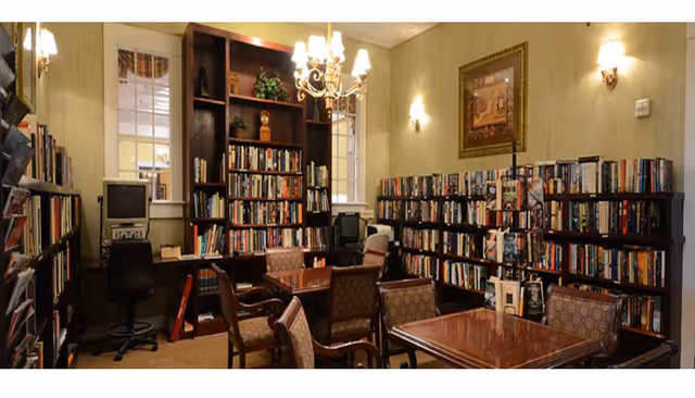 A cozy library room with wooden bookshelves filled with books lining the walls. There are several wooden tables with upholstered chairs arranged for reading or small group gatherings. A chandelier and wall sconces provide warm lighting. A framed painting hangs on the wall, and a computer workstation is set up in one corner.