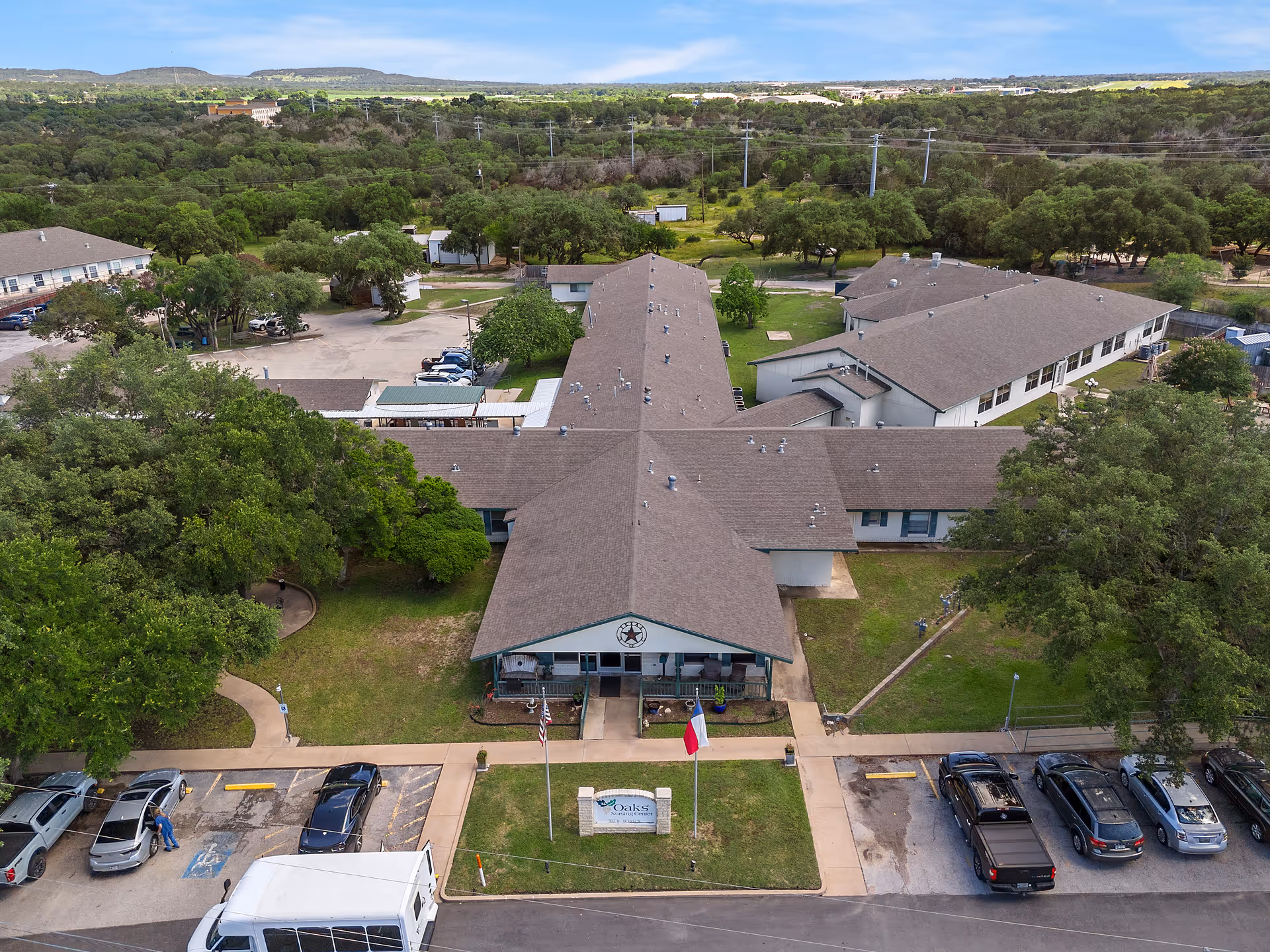 Aerial view of Oaks Nursing Center showing the front entrance and cross-shaped building surrounded by parking, flags, and trees.