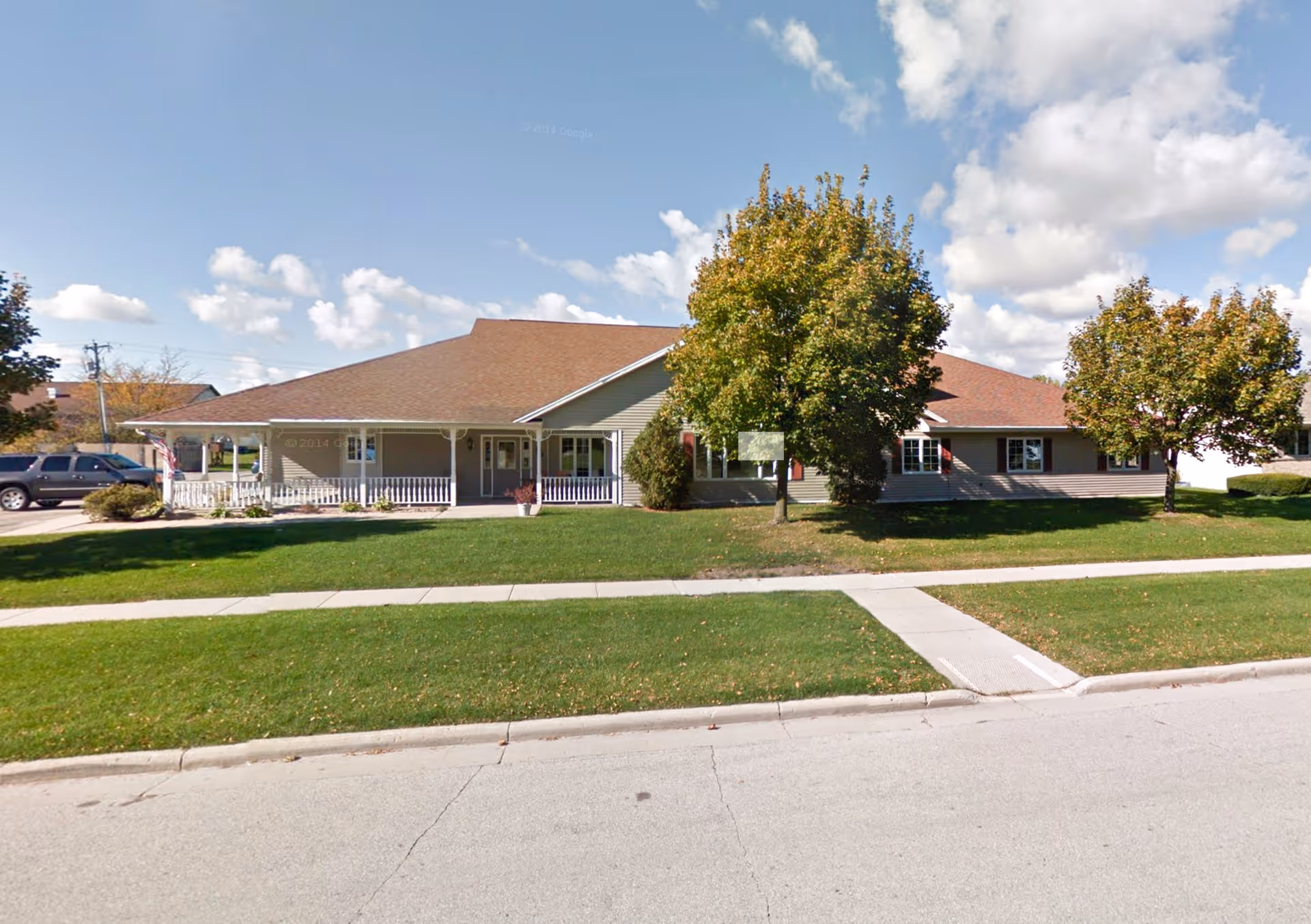 Single-story building with a brown roof and beige siding, surrounded by green grass and trees under a partly cloudy sky. There is a sidewalk in front and a driveway with parked vehicles to the left.