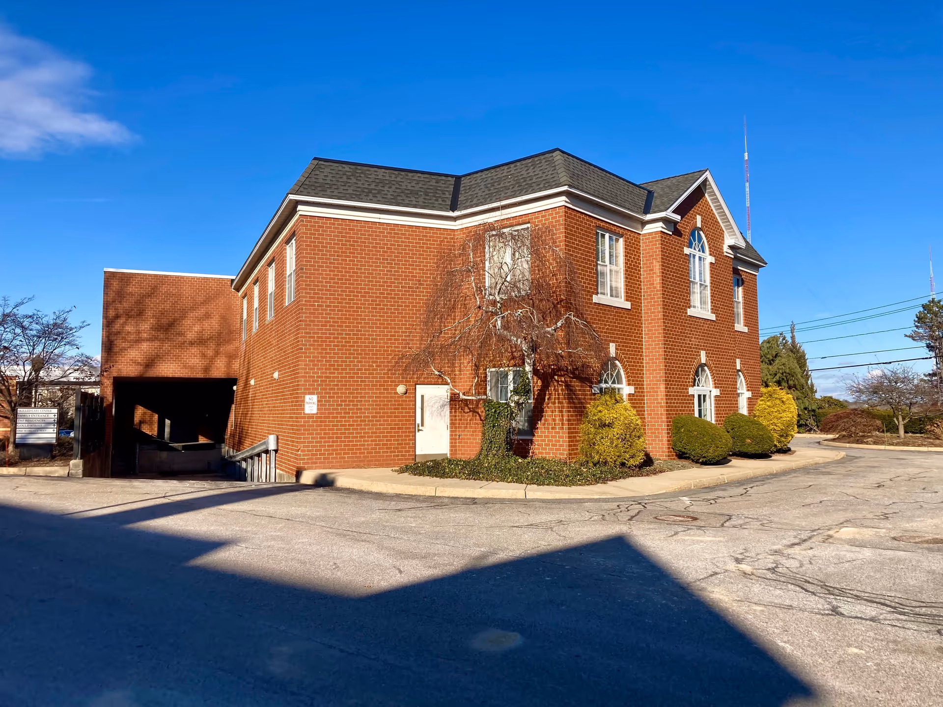 Two-story red brick building with arched windows, shrubs, and a parking area under a clear blue sky.