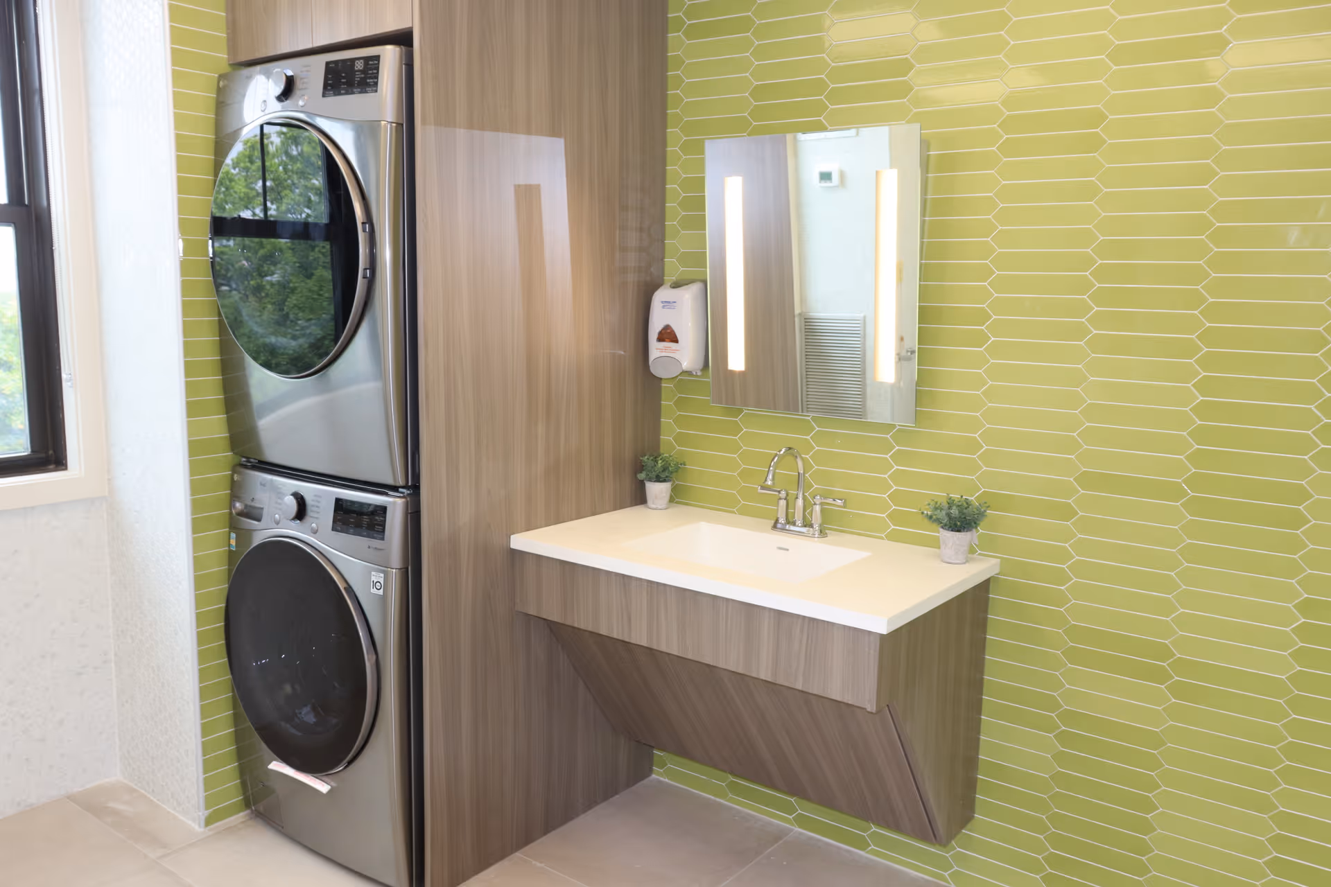 Stacked washer and dryer next to a wall-mounted sink and mirror on a green-tiled interior wall.