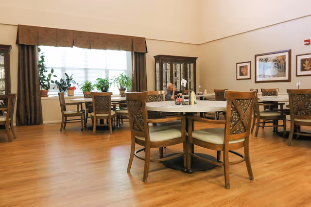 Dining room with round tables and wooden chairs, large windows with potted plants and framed artwork on the walls.