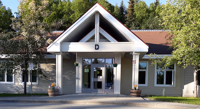 Front entrance of a single-story building with a gabled portico, glass double doors, hanging planters and trees around the walkway.