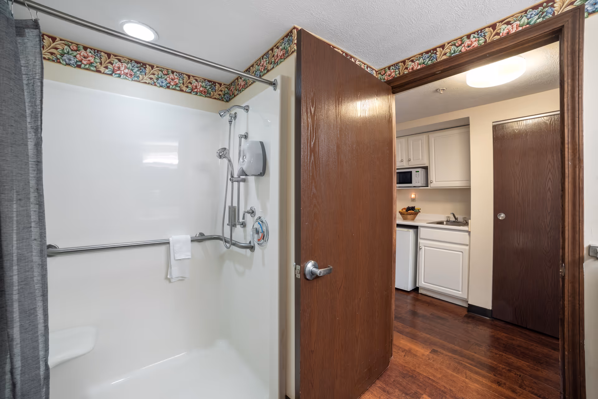 Accessible shower with grab bars and a towel, seen next to an open door revealing a small kitchenette with white cabinets.