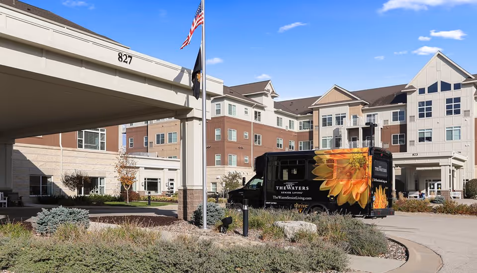 Exterior view of The Waters on Mayowood senior living facility showing a multi-story building with beige and brown siding, an entrance canopy with the number 827, an American flag and another flag on flagpoles, and a black shuttle van with a large yellow flower graphic parked near the entrance.