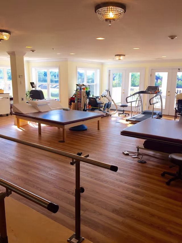 A bright and spacious physical therapy room with wooden floors, parallel bars for walking exercises, two large padded therapy tables, exercise equipment including a treadmill and stationary bike, and large windows letting in natural light.