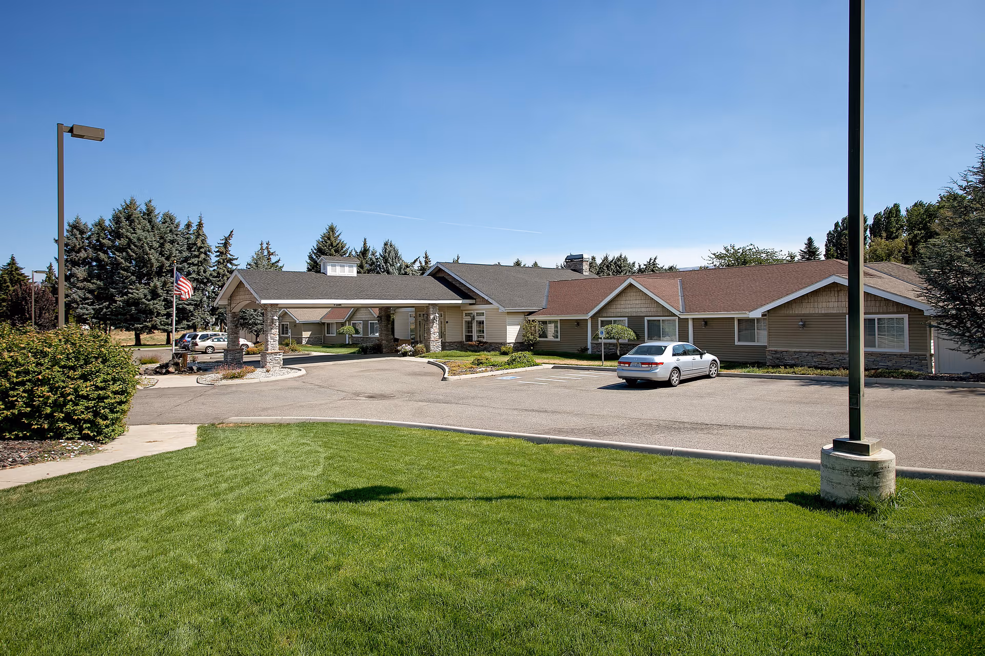 Exterior view of Blossom Creek by Cogir facility showing a single-story building with a covered entrance, a parking area with a silver car, green lawn in the foreground, and trees in the background under a clear blue sky.