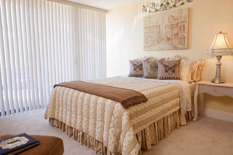 A cozy bedroom featuring a neatly made bed with a beige quilted bedspread and three decorative pillows with butterfly designs. The room has vertical blinds covering a large window, a nightstand with a classic lamp, and a framed artwork on the wall above the bed.
