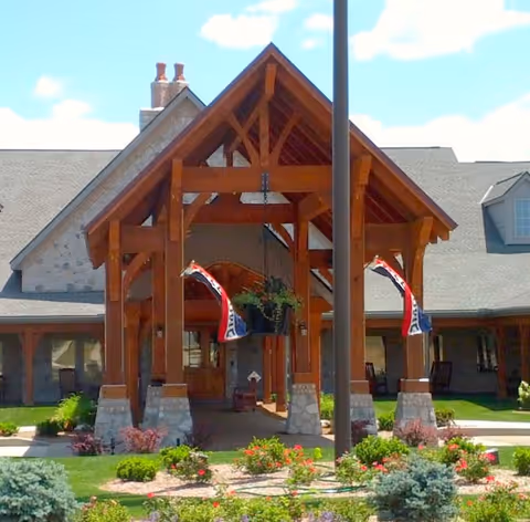 Front entrance of a building with a wooden peaked porch supported by stone pillars, two flags hanging on either side, and a landscaped garden with flowers and shrubs in the foreground under a blue sky with some clouds.