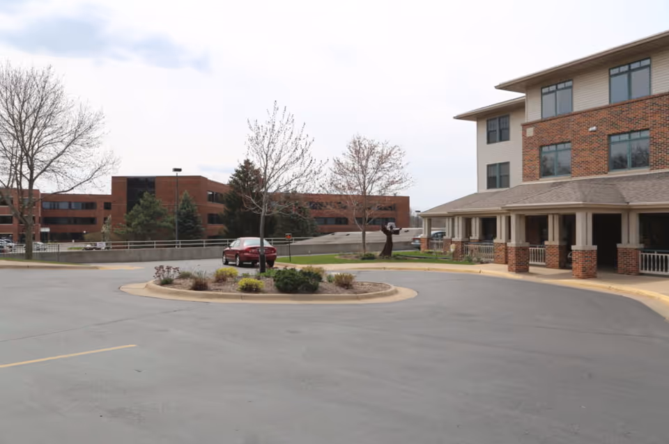 Exterior view of Stonehill Communities Assisi Village showing a circular driveway with a landscaped island in the center, a red car parked near the island, and a multi-story brick and beige building with large windows and a covered entrance. Trees without leaves and another brick building are visible in the background under a cloudy sky.