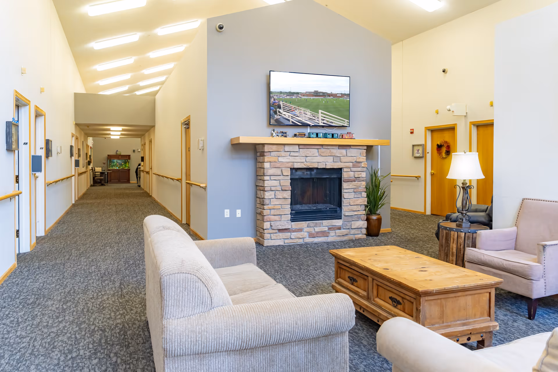 A bright and spacious common area in a senior living facility with a beige couch, armchairs, and a wooden coffee table arranged around a stone fireplace. Above the fireplace is a mounted flat-screen TV displaying a sports game. The room has carpeted floors, light-colored walls, and a long hallway with multiple doors and handrails on both sides. A table lamp and a potted plant are also visible near the fireplace.