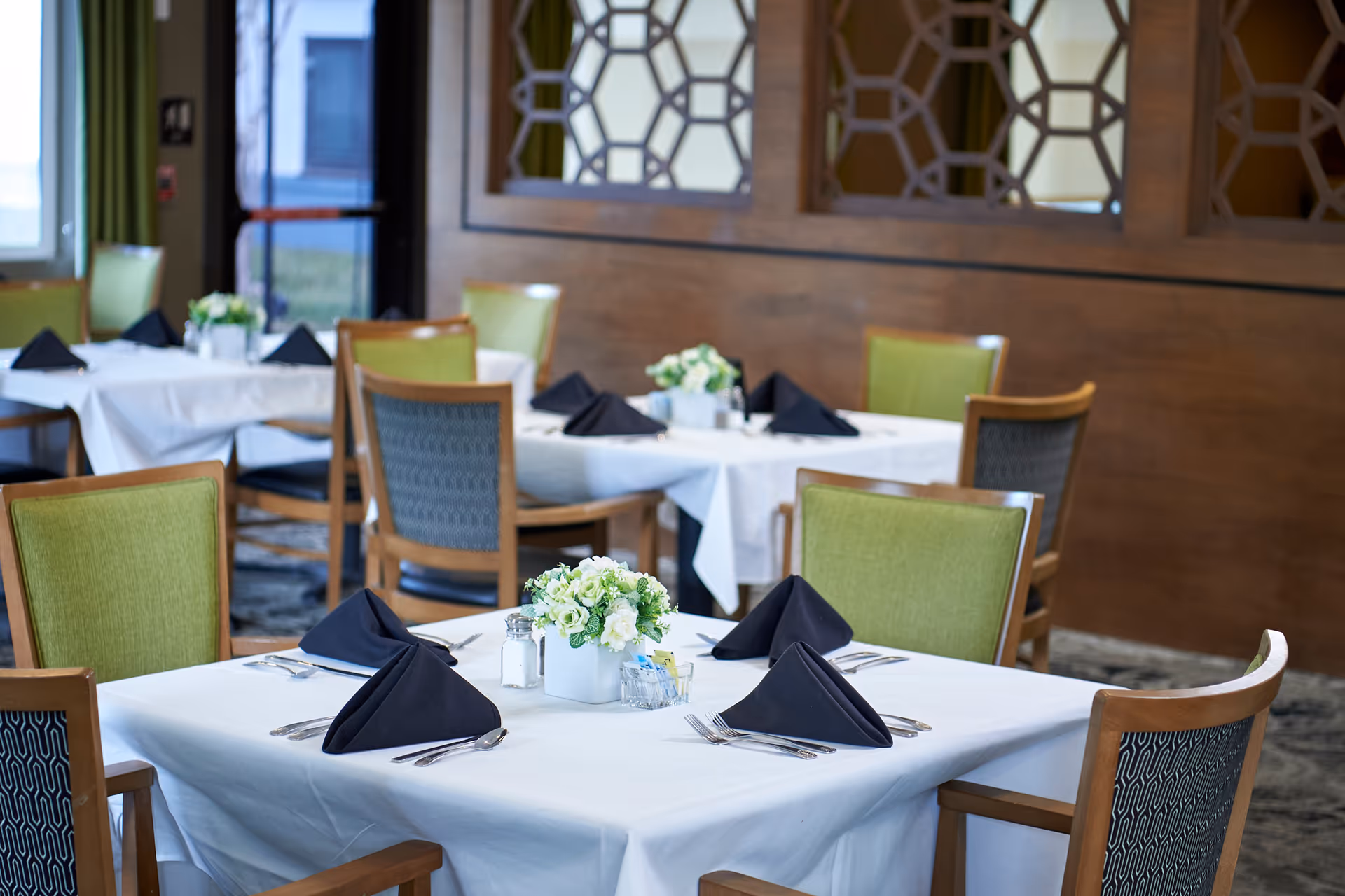 A dining room with tables covered in white tablecloths, each set with black folded napkins, silverware, and small floral centerpieces. The chairs have green and patterned upholstery, and the room features wooden paneling with decorative cutouts on the walls.