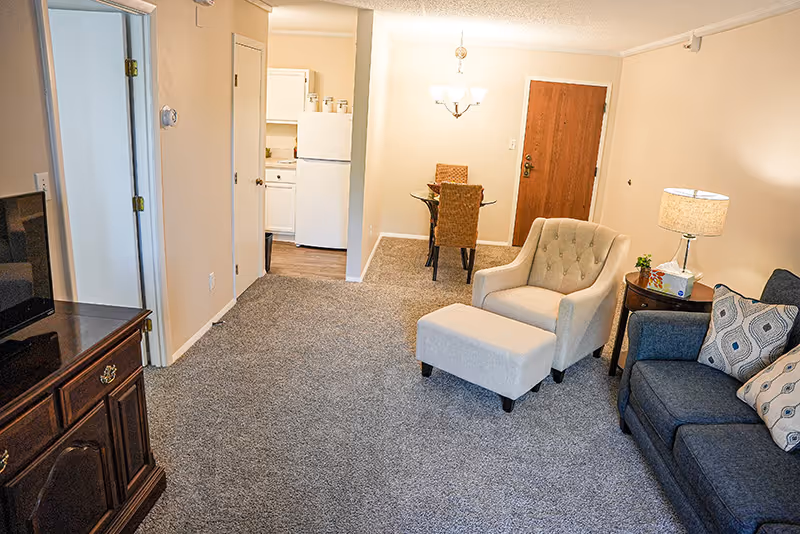 Interior view of a senior living facility apartment showing a living room with a blue couch, a beige armchair with matching ottoman, a wooden side table with a lamp and tissue box, a wooden TV stand with a television, and a dining area with a glass table and two wicker chairs near a wooden door. The kitchen with white cabinets and a refrigerator is visible in the background.