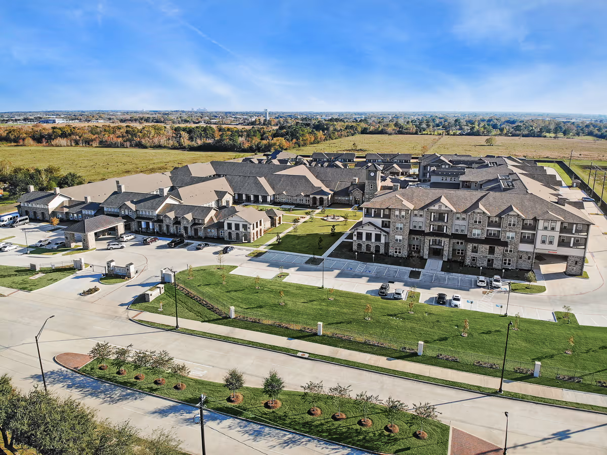 Aerial view of The Lodge at Pine Creek senior living facility showing multiple connected buildings with stone and brick exteriors, surrounded by green lawns, parking lots, and a road in the foreground under a clear blue sky.