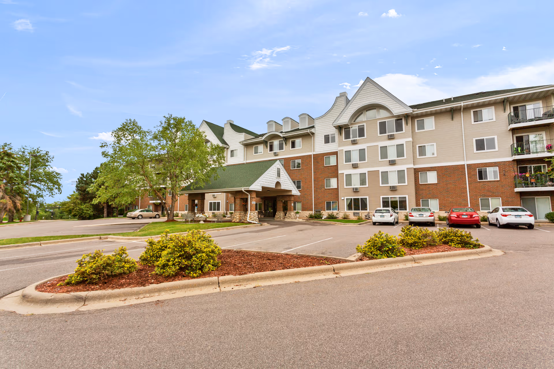 Exterior view of a multi-story senior living facility with a green roof entrance canopy, several parked cars, landscaped bushes, and trees under a partly cloudy sky.