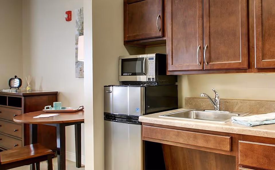 A small kitchen area with wooden cabinets, a stainless steel sink, a mini refrigerator, and a microwave. Adjacent to the kitchen is a wooden dining table with a chair, a cup, and a tray on it. A wooden dresser with a clock and decorative items is visible in the background.