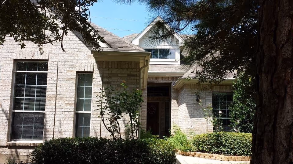 Front exterior view of a brick house with large windows, a peaked roof, and a small garden with bushes and trees in front.