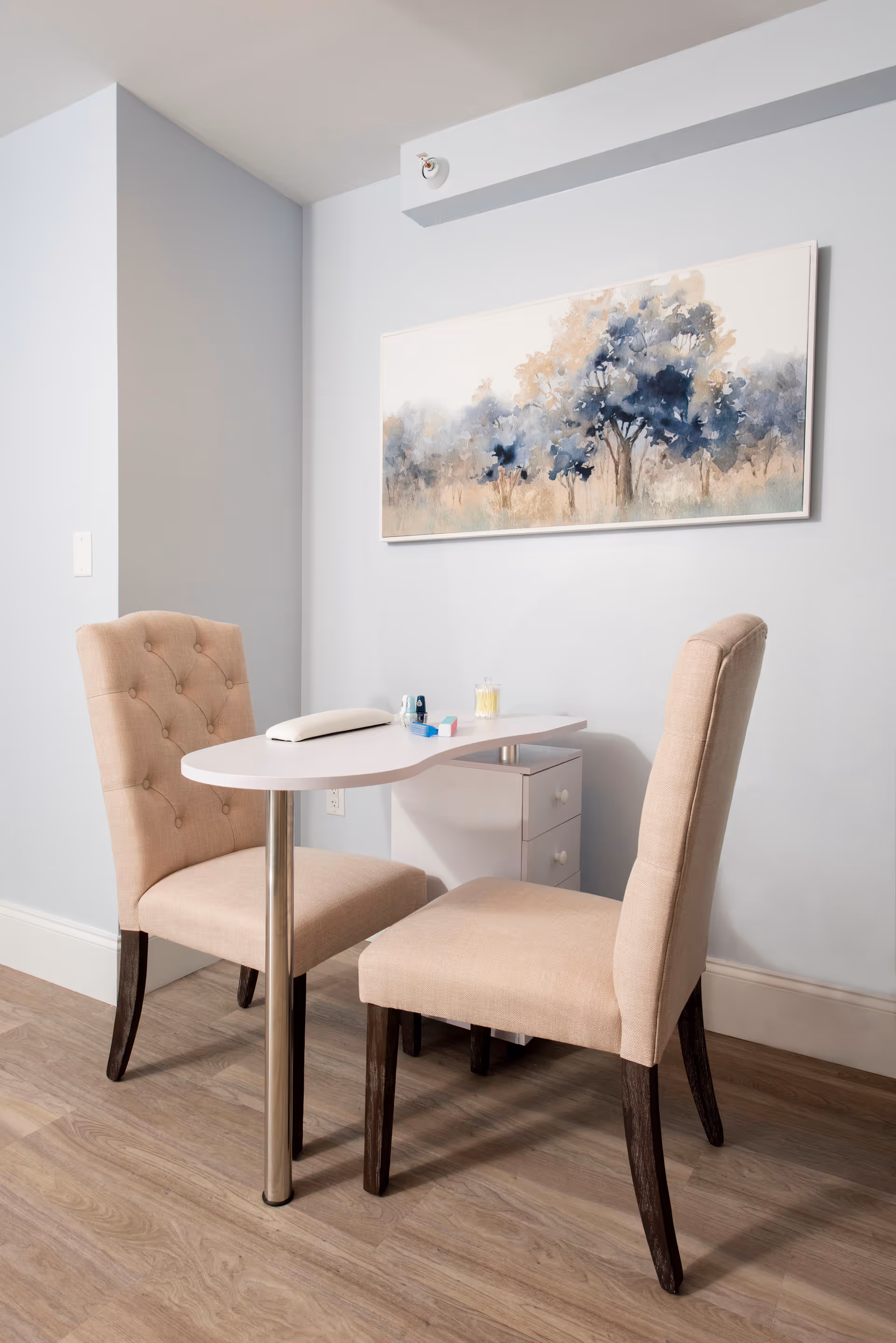 A small white table with a metal leg and a drawer unit underneath, flanked by two beige upholstered chairs with dark wooden legs. On the table are a few small items including a white cushion, a small container with cotton swabs, and some nail polish bottles. A framed painting of trees in soft blue and beige tones hangs on the light gray wall behind the table. The floor is wood with a light finish.