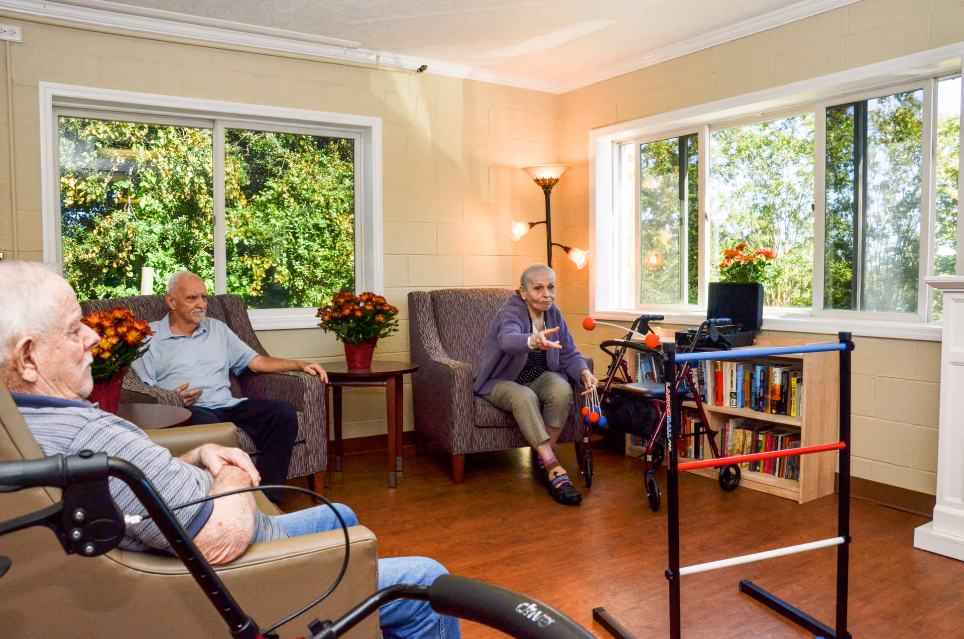 Three elderly individuals sitting in a bright room with large windows showing green trees outside. Two men are seated in armchairs, and a woman is seated in another armchair, actively playing a game involving throwing balls at a ladder-like structure. The room has wooden flooring, a bookshelf, a floor lamp, and potted flowers on side tables.