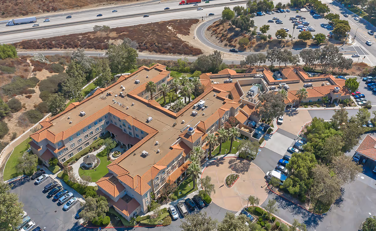 Aerial view of the Bayshire San Dimas senior living complex with terracotta-roofed buildings, circular driveway, parking lots, and nearby highway.