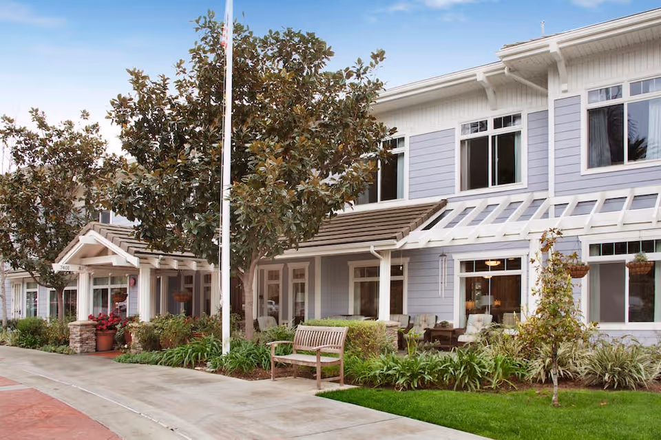 Exterior view of a senior living facility building with light blue siding and white trim. There are large windows, a covered porch area with seating, a wooden bench on the sidewalk, and landscaped greenery including trees and plants. A flagpole is visible in front of the building.