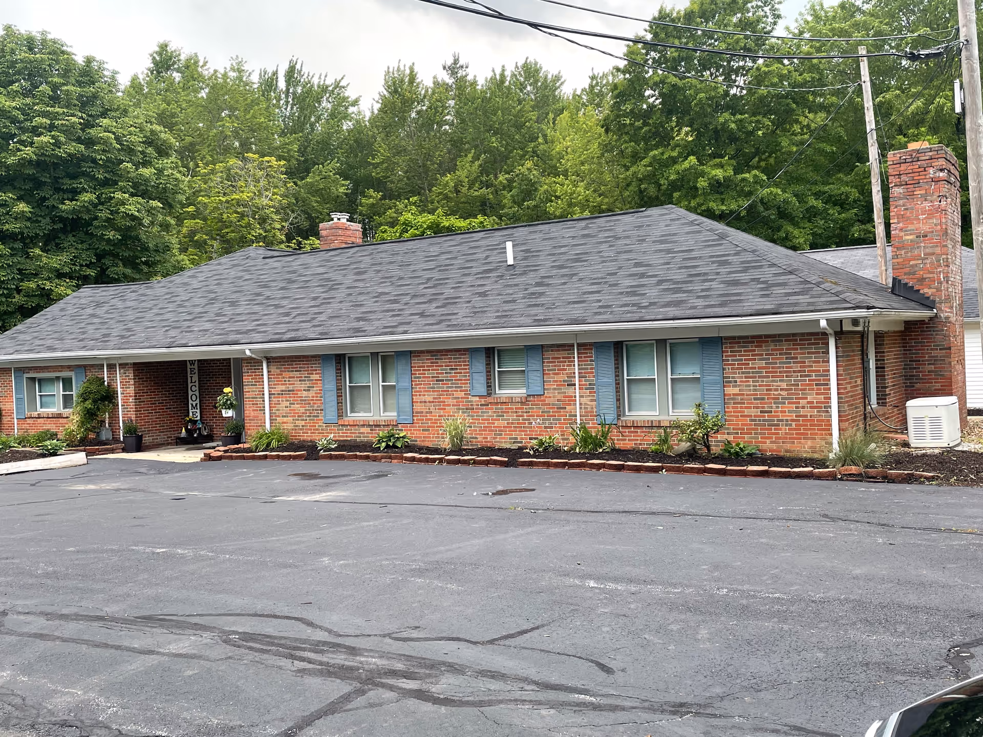 Single-story brick building with a gray shingled roof, blue window shutters, and a chimney. There is a paved parking area in front and greenery including trees and shrubs surrounding the building. A welcome sign is visible near the entrance.