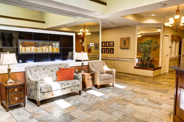 Bright senior living facility lobby with a patterned sofa, armchair, side tables and lamps, tile floor, mirrors and chandeliers.