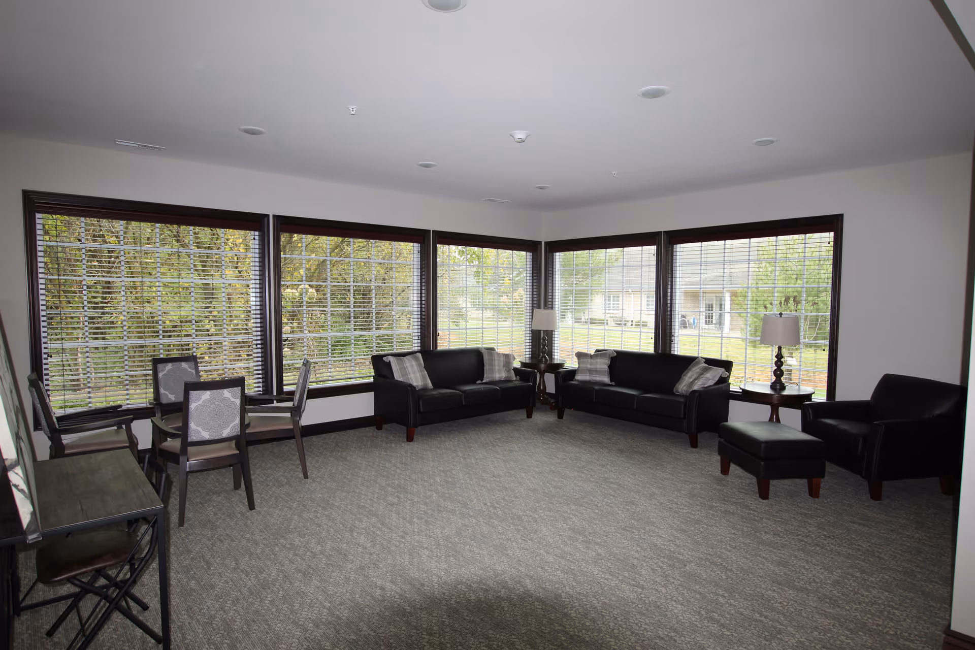 A spacious, well-lit living room with large windows on two walls showing an outdoor view of trees and a building. The room features two black leather sofas with plaid pillows, a black leather armchair with an ottoman, two side tables with lamps, and a small table with four chairs near the windows. The floor is carpeted in a neutral tone.