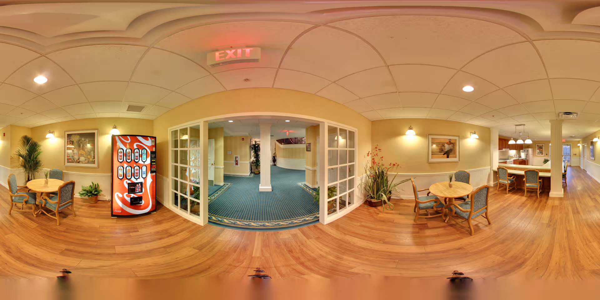 Interior view of a senior living facility common area with wooden floors, round tables with chairs, a Coca-Cola vending machine, and a hallway leading to other rooms. The area is well-lit with ceiling lights and wall sconces, and decorated with framed artwork and plants.
