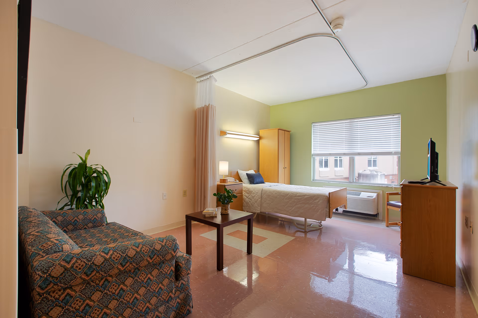 A private senior living room with a single bed, patterned couch, small coffee table, TV, wardrobe and a window.