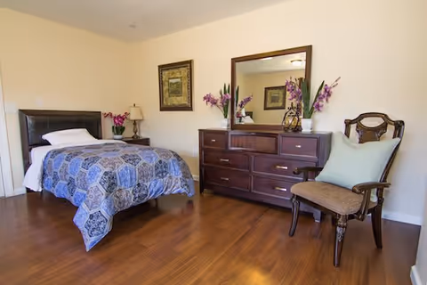 A bedroom with a single bed covered with a blue patterned quilt, a wooden nightstand with a lamp and a vase of flowers, a wooden dresser with a large mirror and decorative flowers, and a wooden chair with a cushion. The room has wooden flooring and light-colored walls.