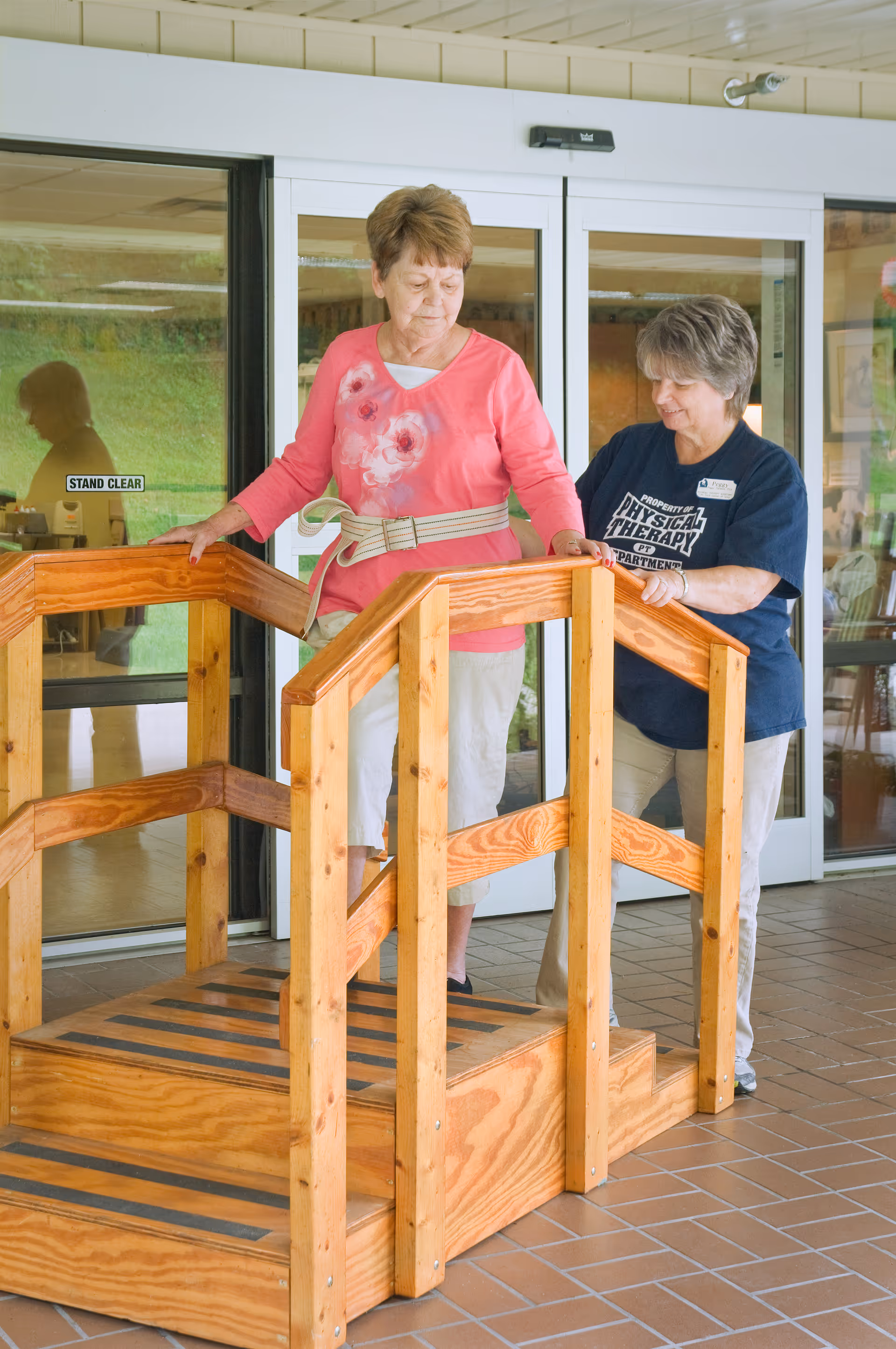 An elderly woman wearing a pink floral shirt and white pants is walking up a small wooden ramp with handrails, assisted by a physical therapist in a navy blue shirt. They are indoors near glass doors with a 'STAND CLEAR' sign.
