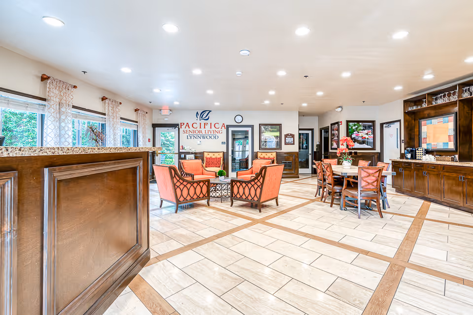 Bright and spacious senior living common area with tiled floor and wooden trim. The room features a seating area with four orange cushioned chairs around a small table, a dining table with six chairs, large windows with patterned curtains, and wooden cabinetry with glassware. The wall displays a sign reading 'Pacifica Senior Living Lynnwood'.