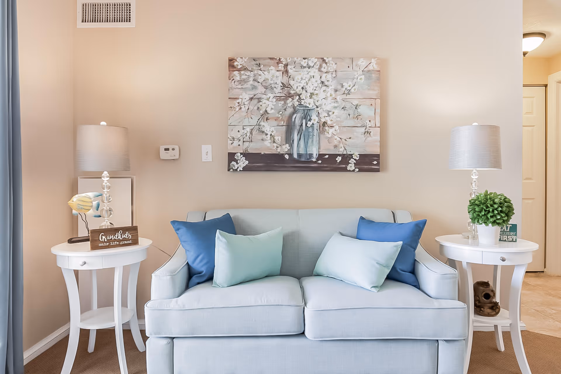 A cozy living room area featuring a light blue loveseat with four cushions in shades of blue and green. On either side of the loveseat are white round side tables, each with a lamp. The left table also has a decorative bird and a sign that reads 'Grandkids make life grand.' The right table has a small green potted plant and a small sign. Above the loveseat hangs a painting of white flowers in a glass jar against a rustic wooden background. The walls are painted beige, and there is a blue curtain partially visible on the left side.