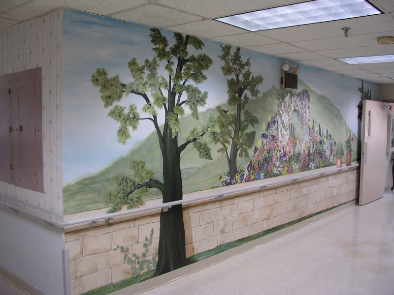 A hallway with a mural painted on the wall depicting trees, flowers, and a scenic landscape with hills in the background. The floor is tiled, and there is a door open at the end of the hallway.