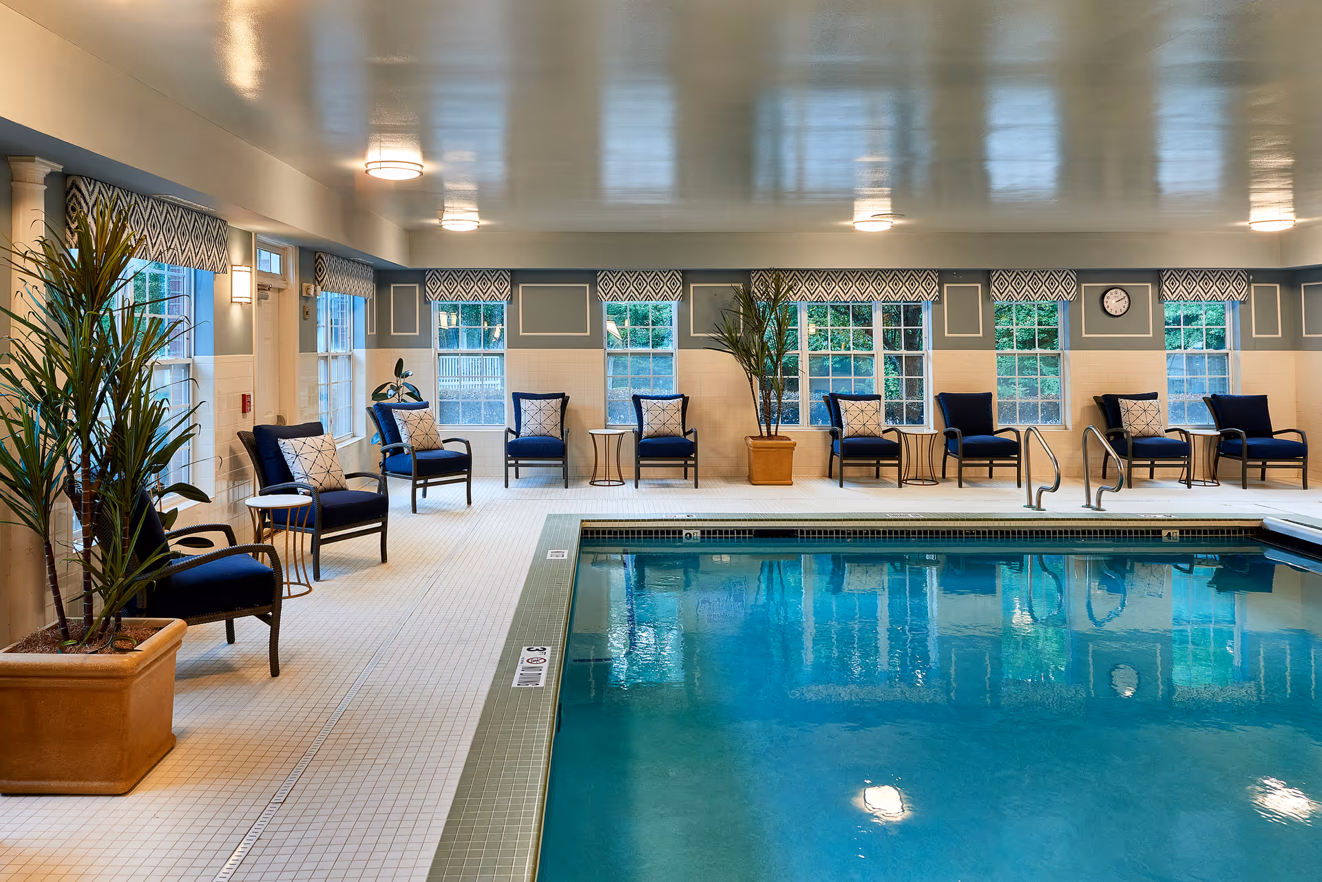 Indoor swimming pool area with clear blue water, surrounded by a tiled floor. Several navy blue cushioned chairs with patterned pillows are arranged along the walls near large windows with geometric valances. There are potted plants placed near the seating area, and ceiling lights illuminate the space.